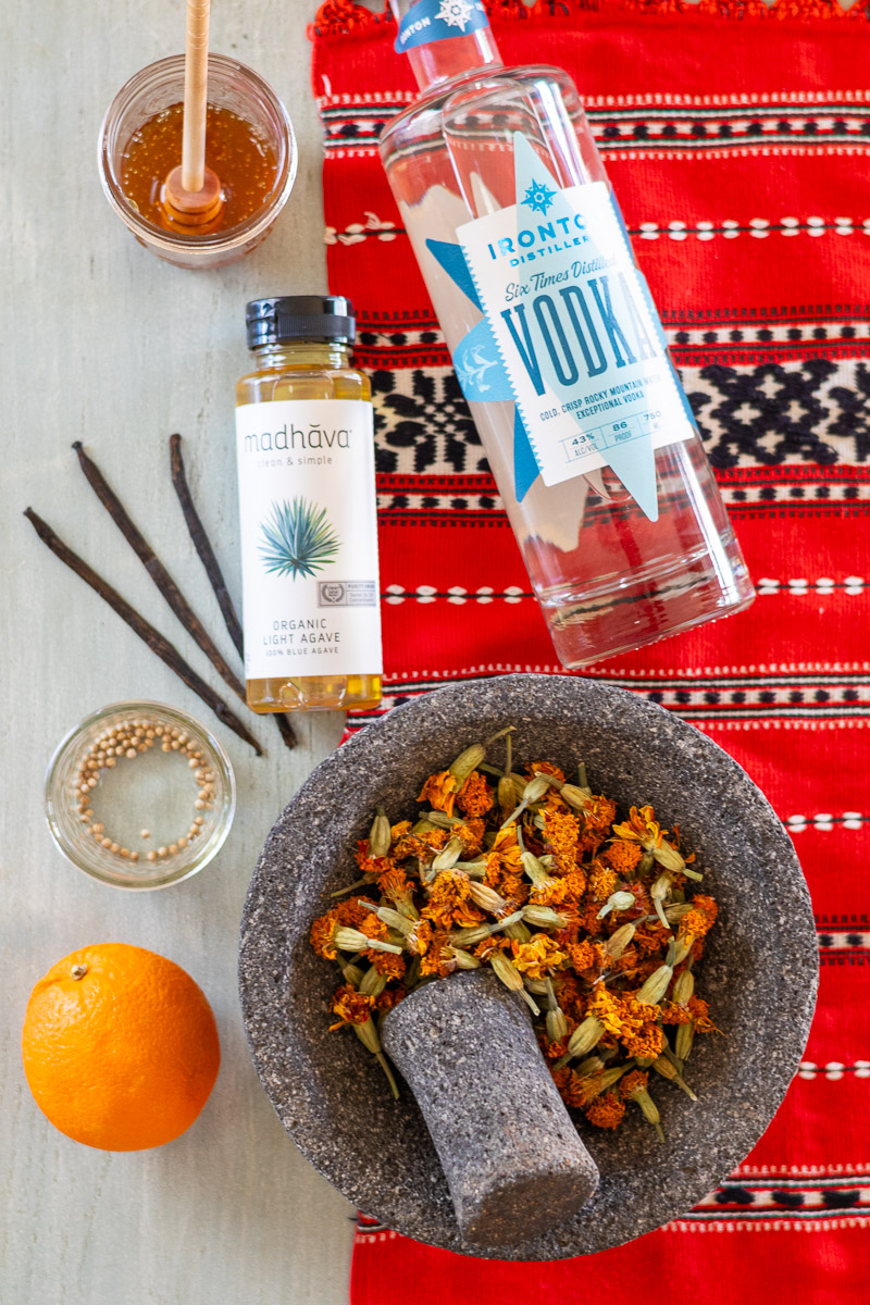 Honey, vodka, agave, vanilla beans, coriander, marigold blossoms in mortar and pestle, and orange laid out on a table under a red table runner.