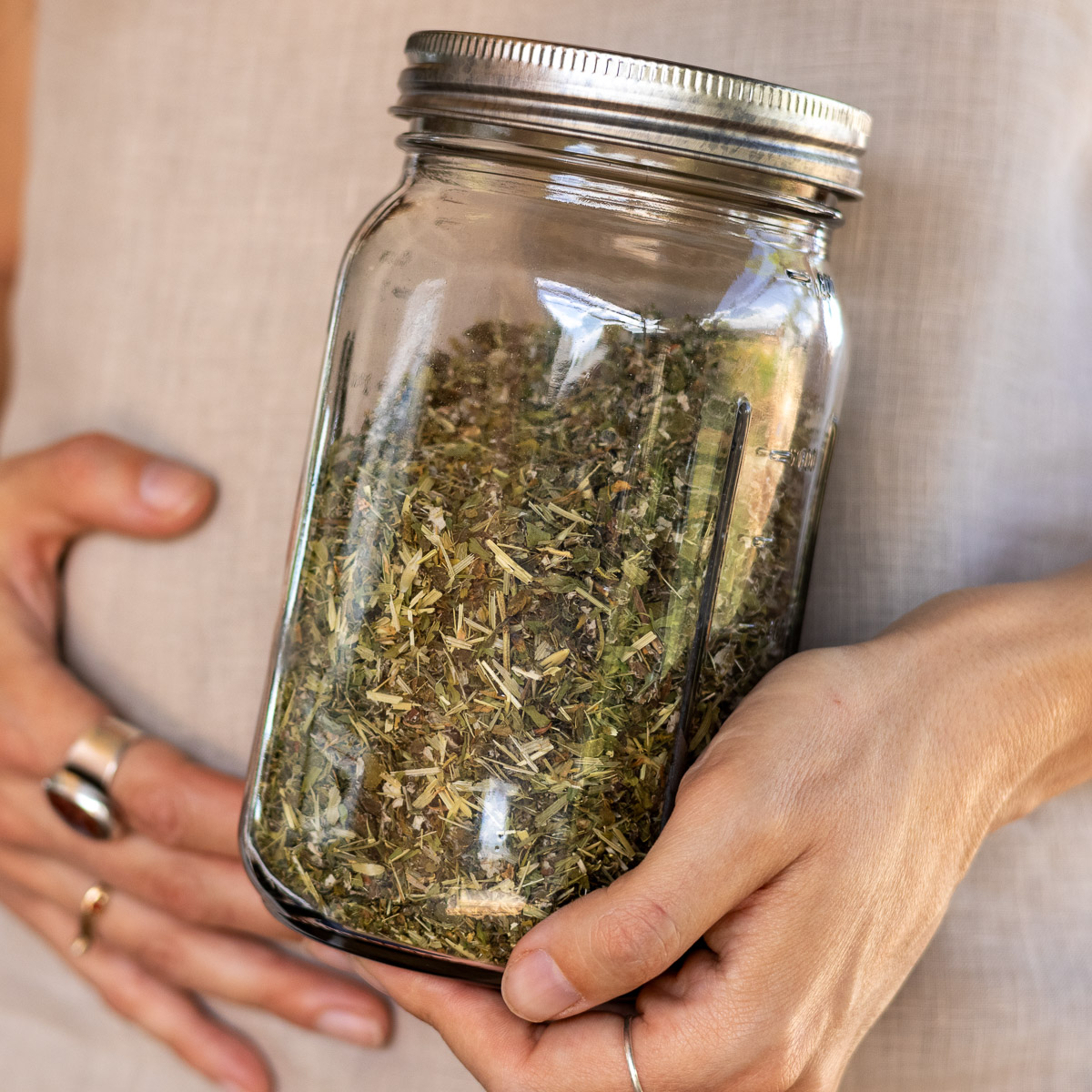 A mason jar of blended pregnancy tea blend and a woman in the background holding her belly with a hand with rings.